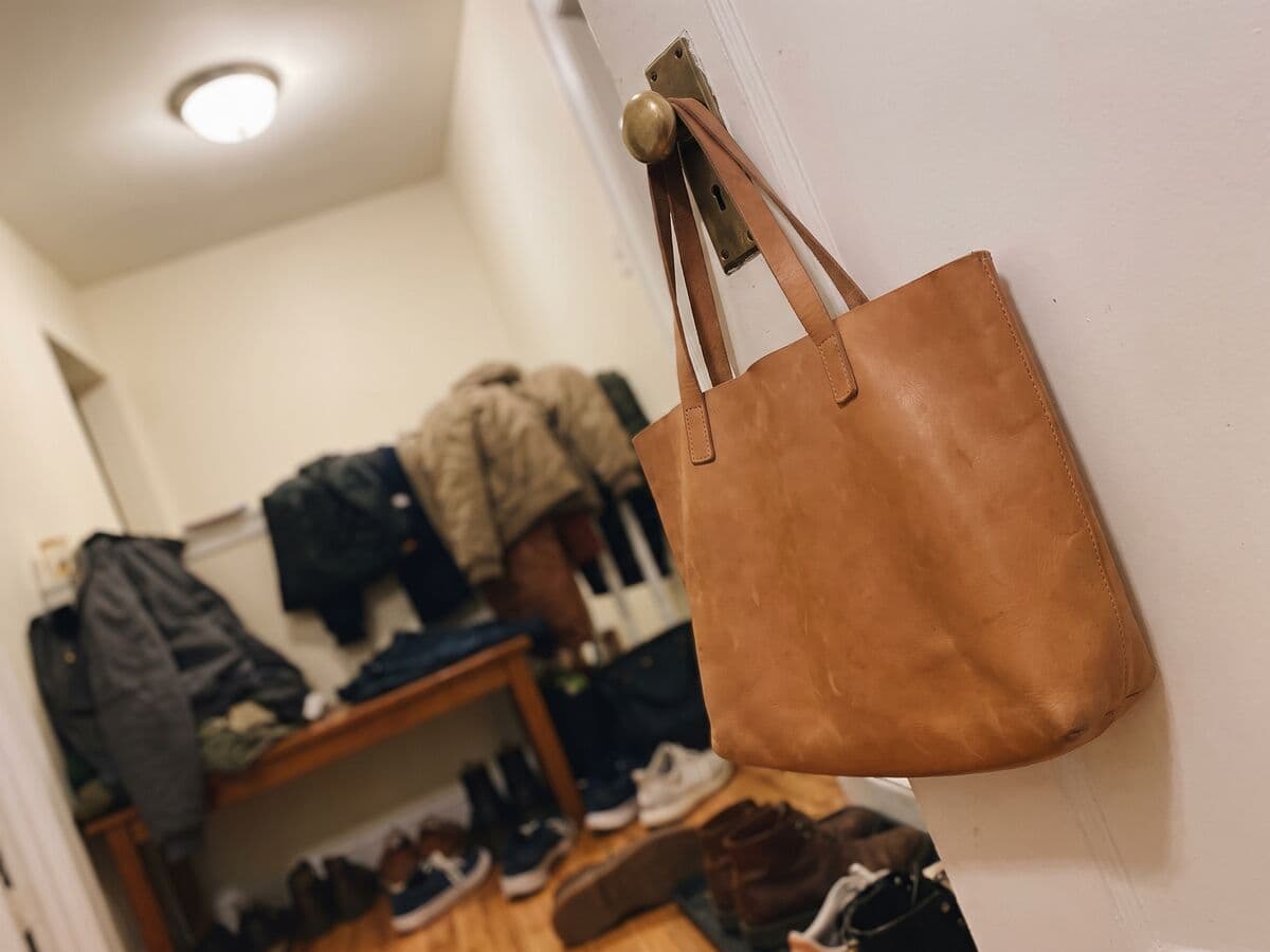 A tan leather tote bag hanging on a doorknob in a cluttered hallway