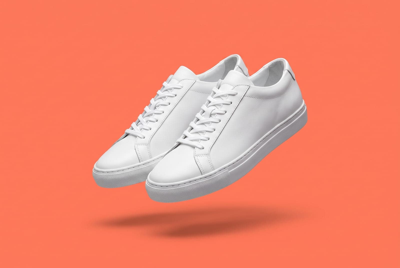A pair of white leather sneakers cut out and placed on a flat coral background with crisp edges