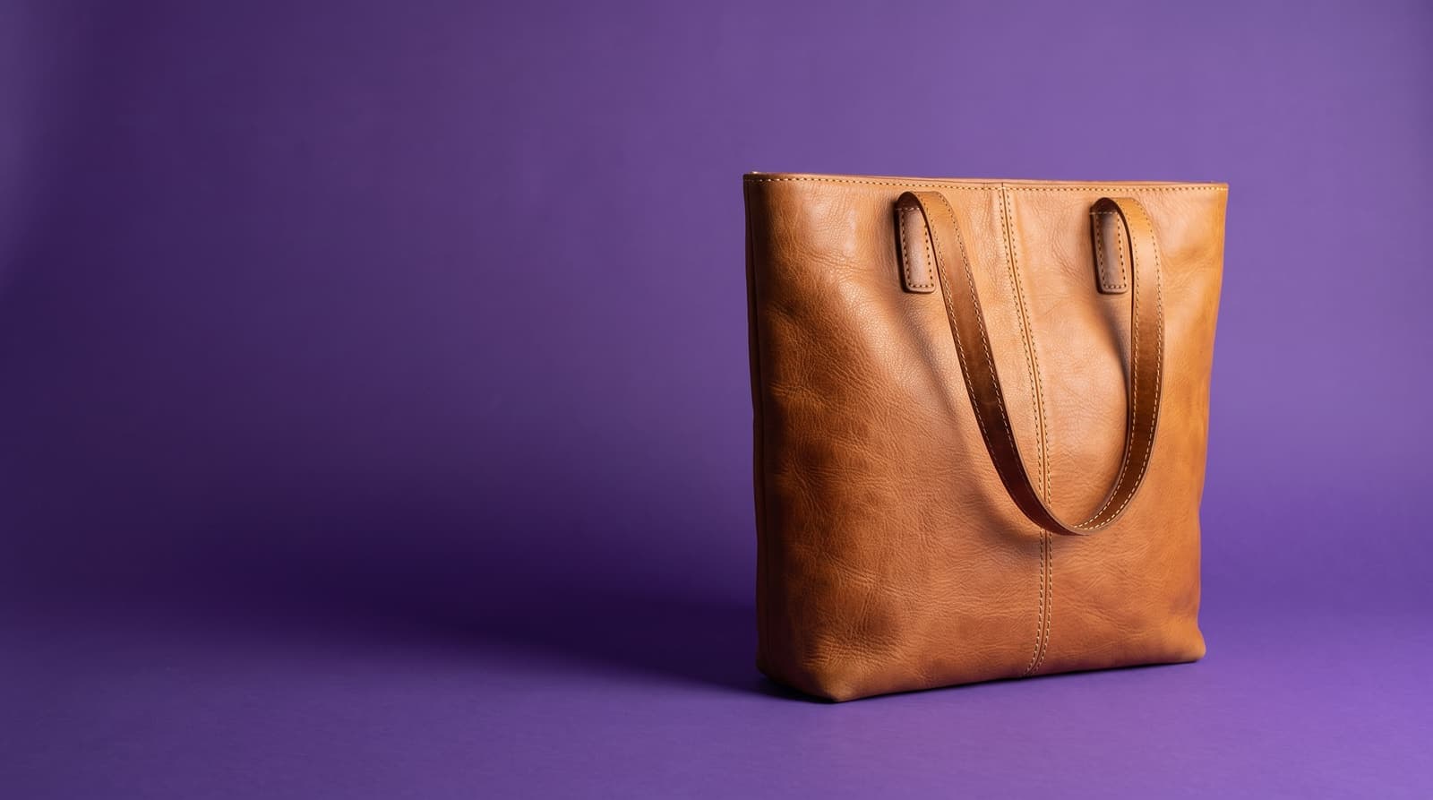A tan leather tote bag on a flat purple background with a soft shadow