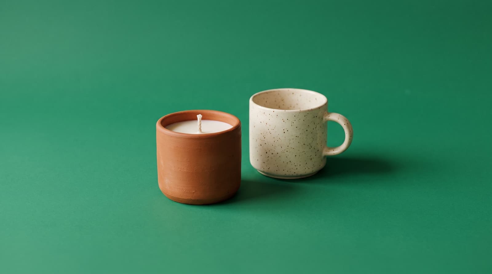 A terracotta candle and a speckled cream mug on a flat emerald green background